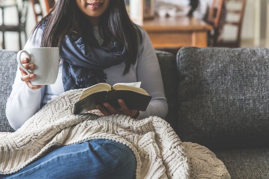 Woman sitting on a cozy gray couch, wrapped in a cream knit blanket, holding a white mug and reading an open Bible, creating a peaceful and reflective atmosphere.