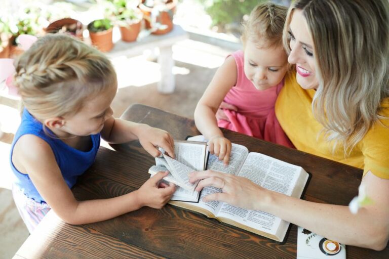 A mother in a mustard-colored top sits at a wooden table with her two young daughters, reading and discussing the Bible together. One child, in a blue dress, eagerly turns a page while the other, in pink, sits on her mother’s lap. Sunlight filters through an outdoor setting with potted plants in the background. A warm, joyful moment of teaching and learning God’s Word, reflecting the theme of walking in wisdom in everyday life.