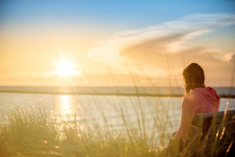 A woman sits on a bench overlooking the water at sunrise, reflecting on the beauty of the moment. The warm golden light highlights the peaceful setting, evoking a sense of contemplation and focus on what truly matters.