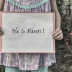 A young girl in a pastel dress holding a sign that reads ‘He is Risen!’ outdoors with blooming flowers in the background, symbolizing the joy and hope of Christ’s resurrection on Easter.