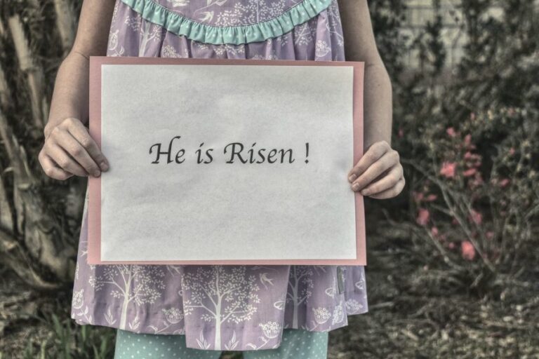 A young girl in a pastel dress holding a sign that reads ‘He is Risen!’ outdoors with blooming flowers in the background, symbolizing the joy and hope of Christ’s resurrection on Easter.