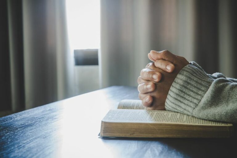 Hands clasped in prayer over an open Bible on a wooden table, with soft natural light coming through a window—symbolizing humility, reflection, and seeking God’s wisdom over self-promotion.