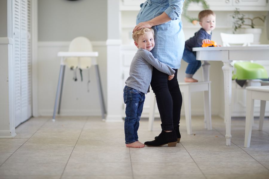 Young boy hugging his mother in a bright, peaceful kitchen while another child plays at the table—capturing a life-giving moment of love and connection in everyday homemaking.
