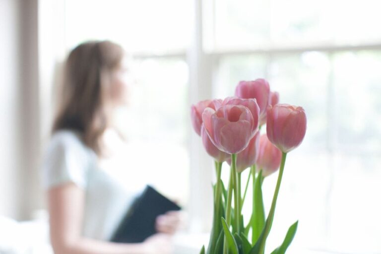 A soft image of pink tulips in focus with a woman holding a Bible and gazing out the window in the background, symbolizing quiet reflection and finding joy in Christ-centered self-forgetfulness.