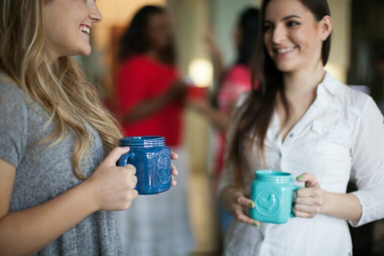 Two women smiling and holding coffee mugs while enjoying conversation, symbolizing friendship, trust, and the importance of believing the best in others.