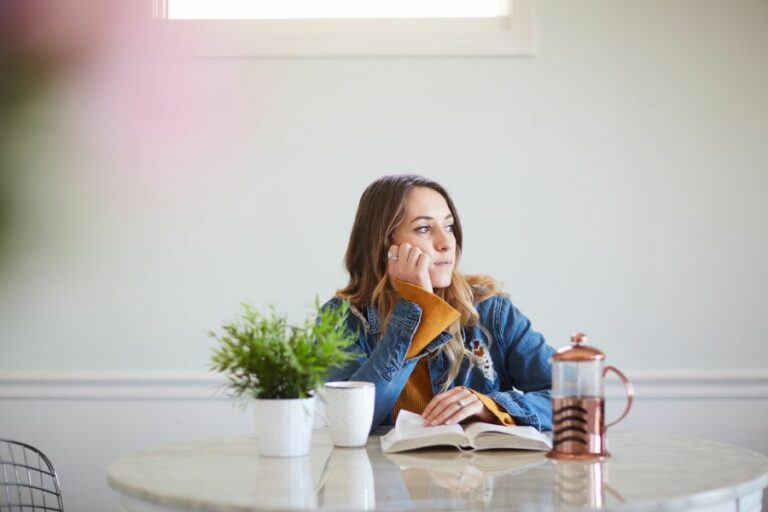 "Woman sitting at a table with an open book and coffee, gazing thoughtfully out of a window, reflecting in a peaceful, light-filled room."